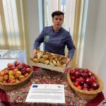 postać trzymająca koszyk z pieczywem stojąca przy stole z koszami jabłek