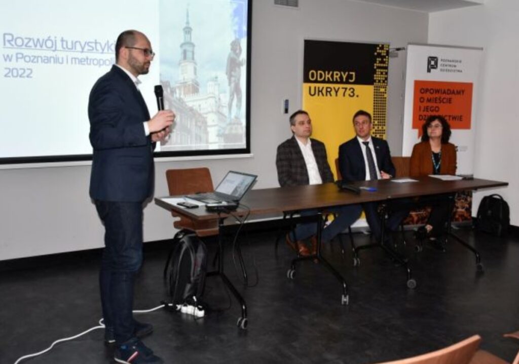 Prelegent na tle ekranu multimedialnego oraz w dalszym planie trójka pozostałych prowadzących