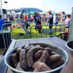 na peirwszym planie miska białych kiełbas, na drugim uczestnicy pikniku plenerowego